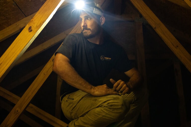 Termite control technician inspecting an attic with a headlamp, checking for termites and other pests in a wooden structure.